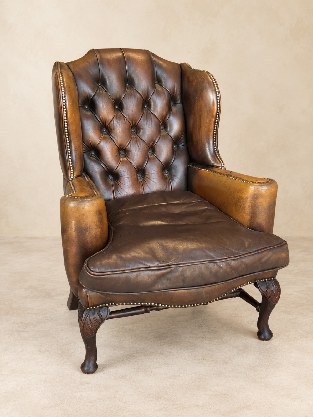 A deep buttoned tan leather wingback armchair featuring brass stud trim, contoured wings, and carved wooden cabriole front legs.