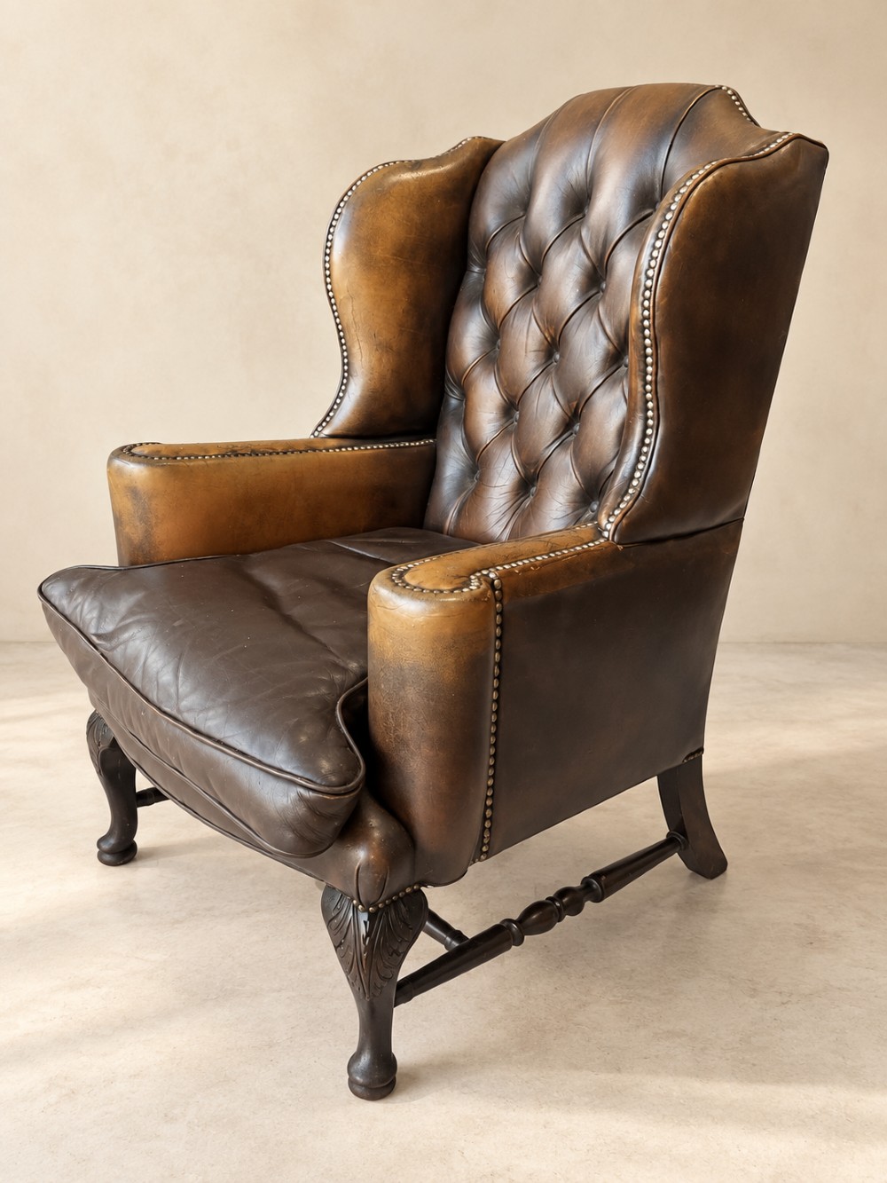 A deep buttoned tan leather wingback armchair featuring brass stud trim, contoured wings, and carved wooden cabriole front legs. View II