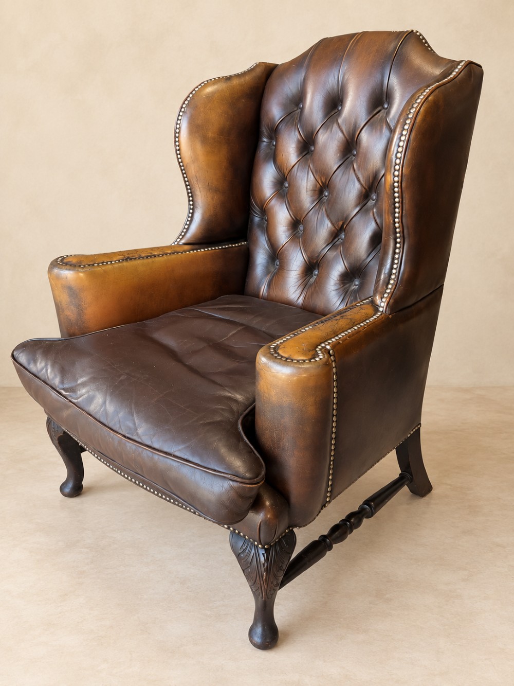 A deep buttoned tan leather wingback armchair featuring brass stud trim, contoured wings, and carved wooden cabriole front legs. View III