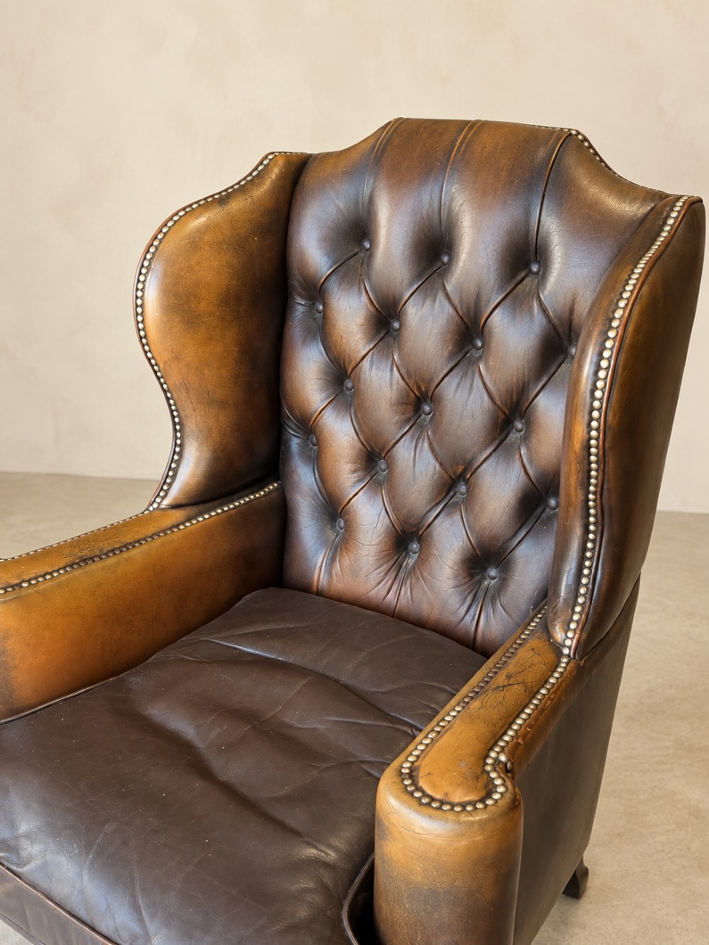 A deep buttoned tan leather wingback armchair featuring brass stud trim, contoured wings, and carved wooden cabriole front legs. View IV