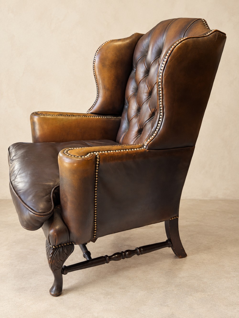 A deep buttoned tan leather wingback armchair featuring brass stud trim, contoured wings, and carved wooden cabriole front legs. View V