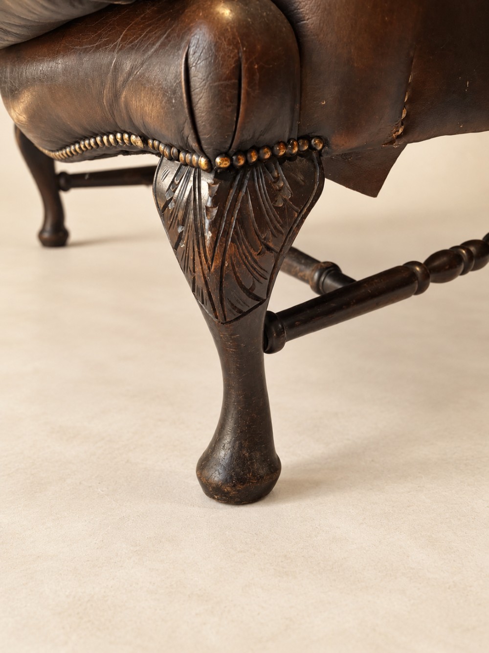 A deep buttoned tan leather wingback armchair featuring brass stud trim, contoured wings, and carved wooden cabriole front legs. View VI