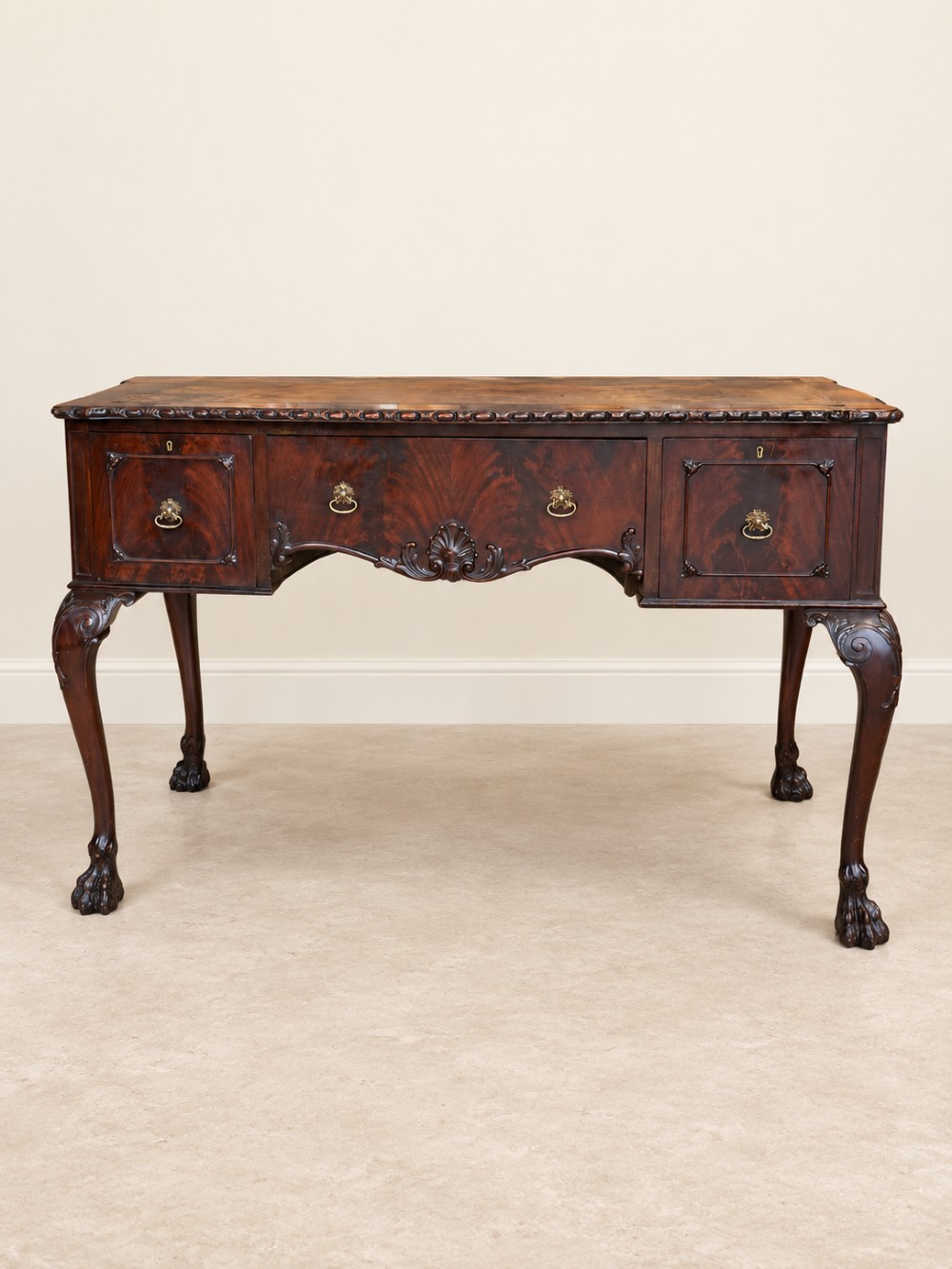 A dark mahogany writing table featuring three drawers, a carved shell apron, and cabriole legs with ball-and-claw feet.