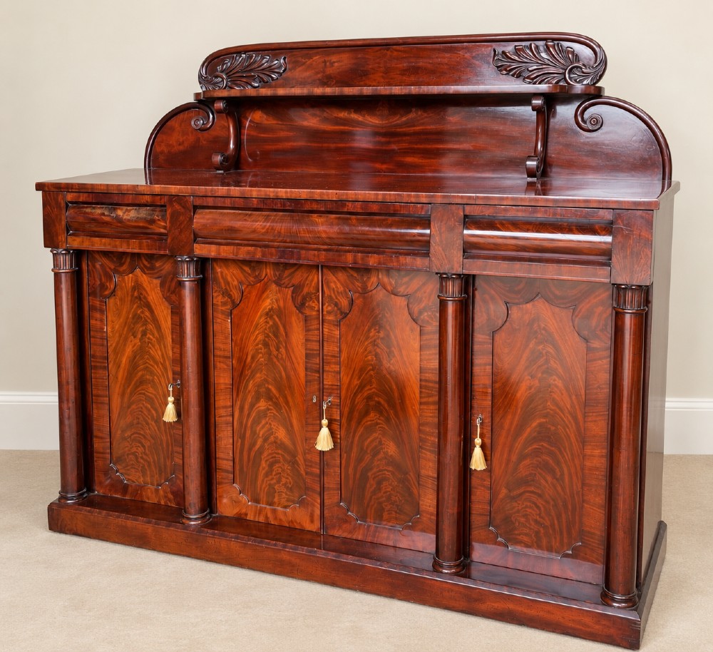 A 19th-century mahogany sideboard with a carved scrolled backboard, flame mahogany door panels, fluted columns, and a solid plinth base.