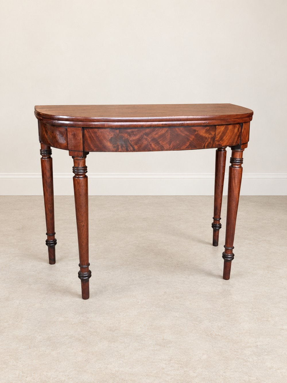 A Regency period mahogany fold-over table featuring a demilune top, flame mahogany frieze, and turned tapered legs with ring detailing. View II