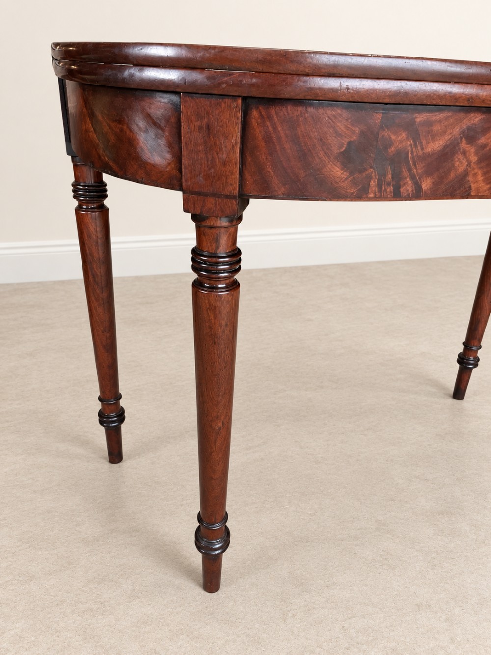 A Regency period mahogany fold-over table featuring a demilune top, flame mahogany frieze, and turned tapered legs with ring detailing. View IV