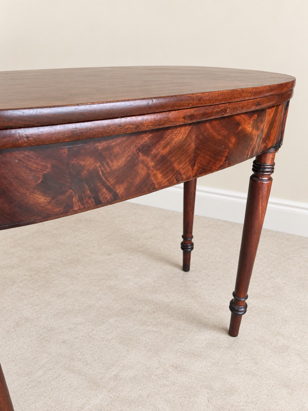 A Regency period mahogany fold-over table featuring a demilune top, flame mahogany frieze, and turned tapered legs with ring detailing. View V