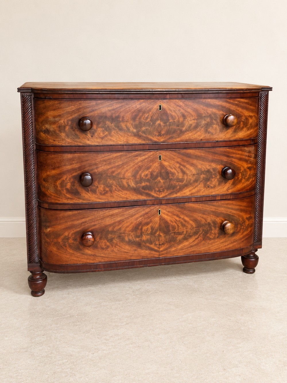 A Regency mahogany chest of drawers featuring a bowfront silhouette, three flame-veneered drawers with turned wooden handles, and reeded side columns.