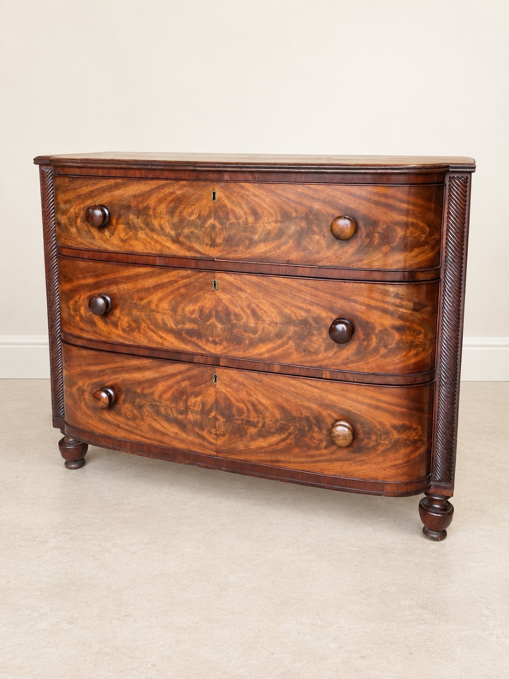 A Regency mahogany chest of drawers featuring a bowfront silhouette, three flame-veneered drawers with turned wooden handles, and reeded side columns. View II