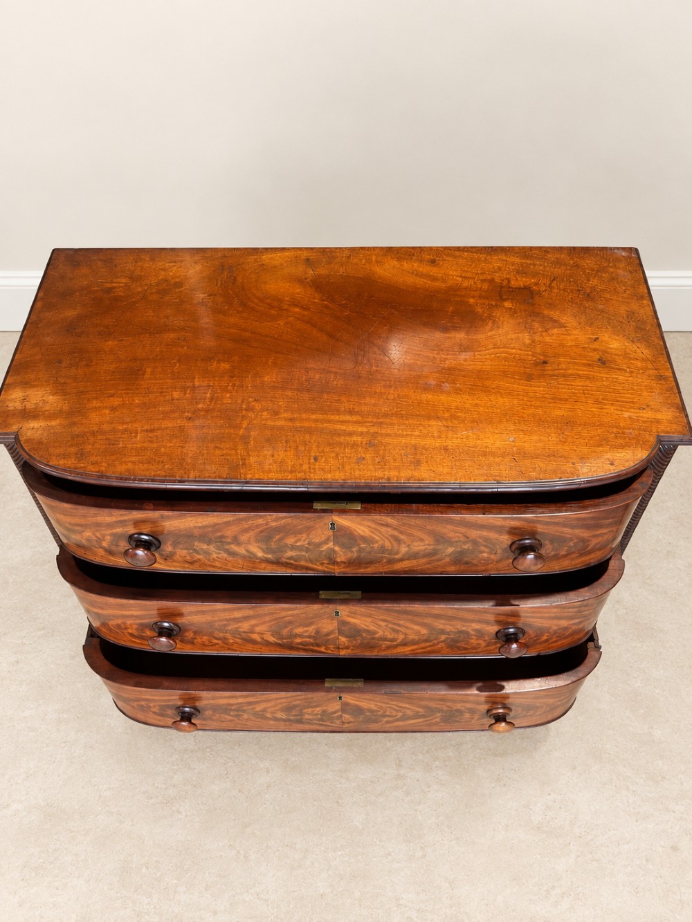 A Regency mahogany chest of drawers featuring a bowfront silhouette, three flame-veneered drawers with turned wooden handles, and reeded side columns. View V