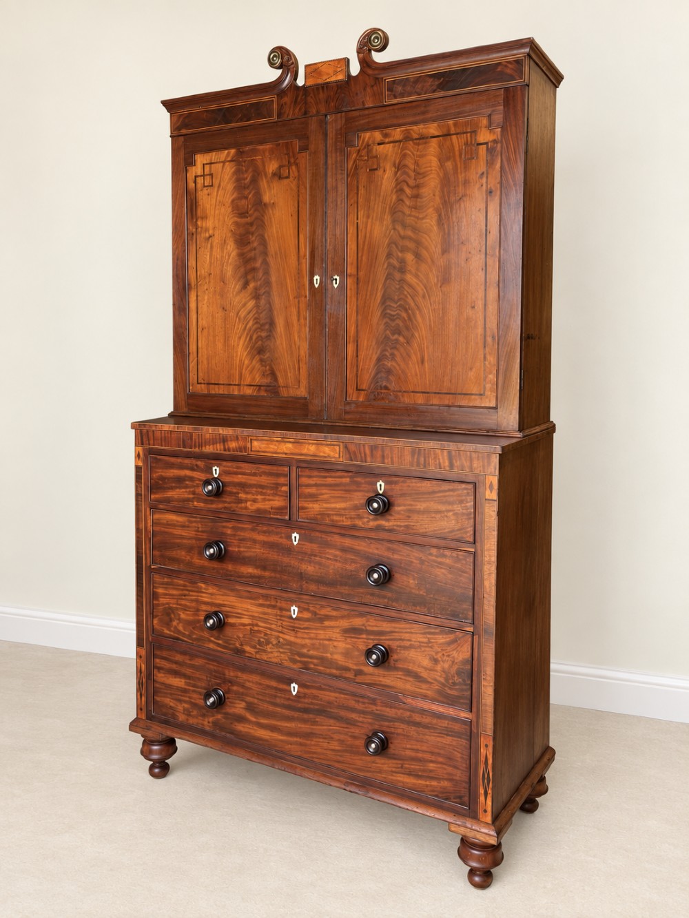 A Georgian mahogany cupboard on chest featuring upper double panelled doors with flame veneer, decorative cresting, and a lower chest of drawers on turned feet.