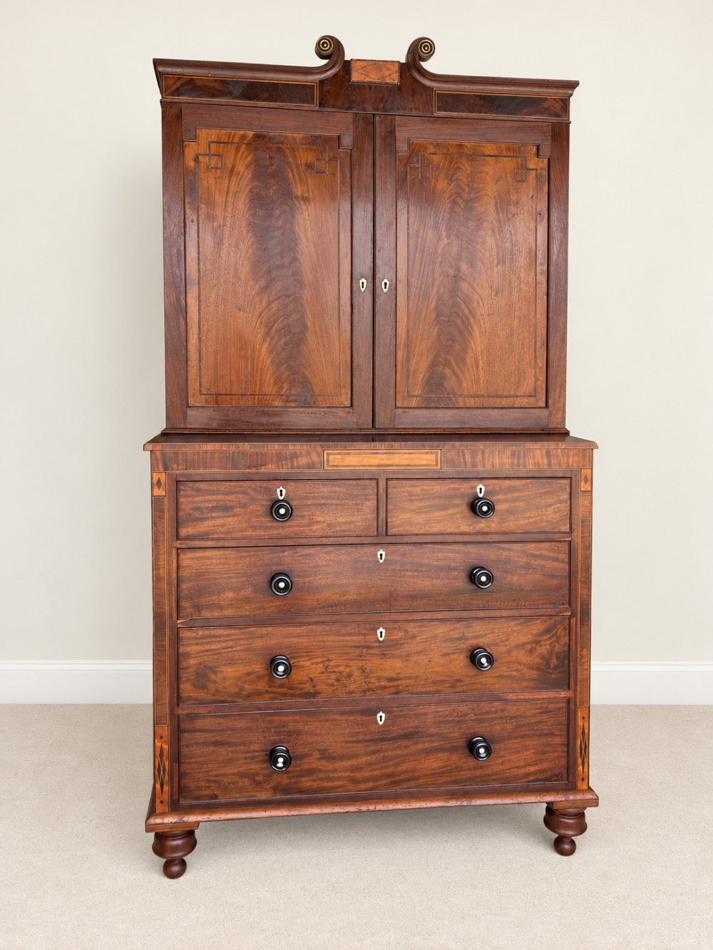 A Georgian mahogany cupboard on chest featuring upper double panelled doors with flame veneer, decorative cresting, and a lower chest of drawers on turned feet. View II