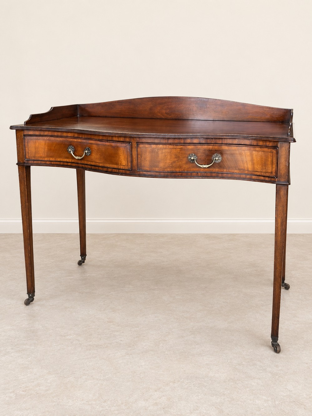 A Georgian style mahogany side table featuring a curved front apron, two drawers with brass swan-neck handles, a low shaped gallery back, and square tapered legs on castors. View II