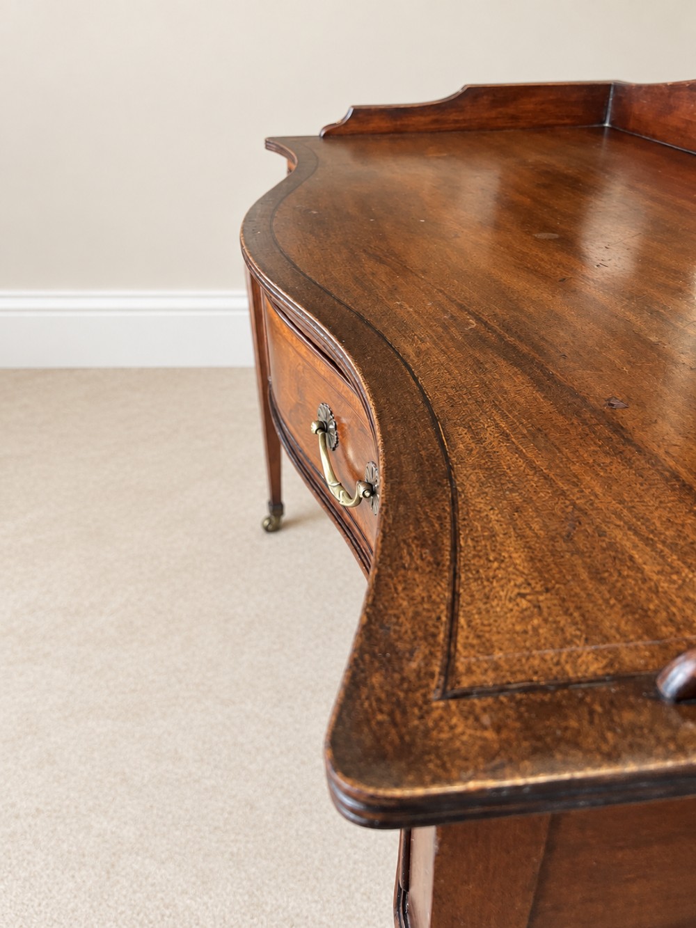 A Georgian style mahogany side table featuring a curved front apron, two drawers with brass swan-neck handles, a low shaped gallery back, and square tapered legs on castors. View IV
