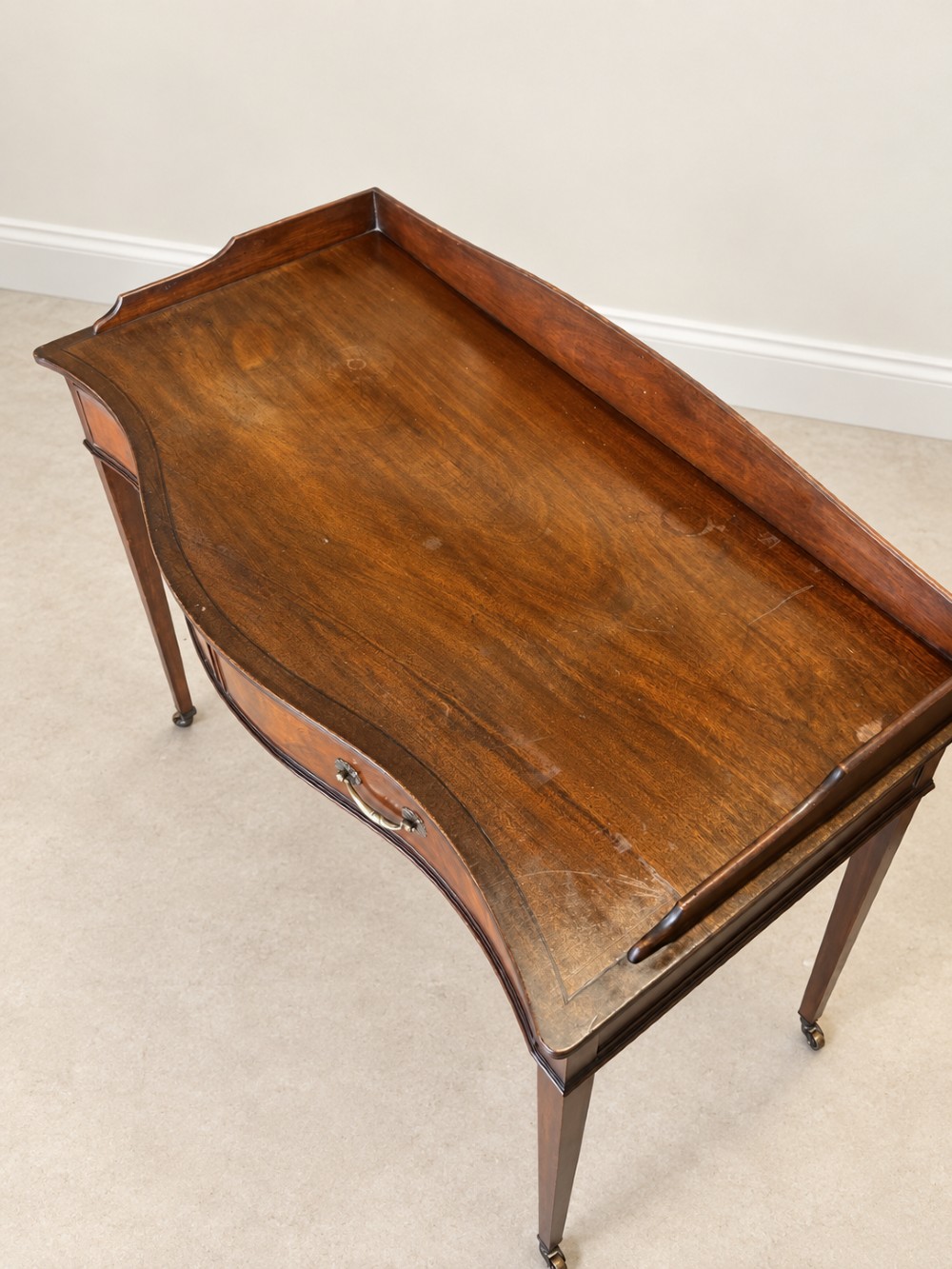 A Georgian style mahogany side table featuring a curved front apron, two drawers with brass swan-neck handles, a low shaped gallery back, and square tapered legs on castors. View VI