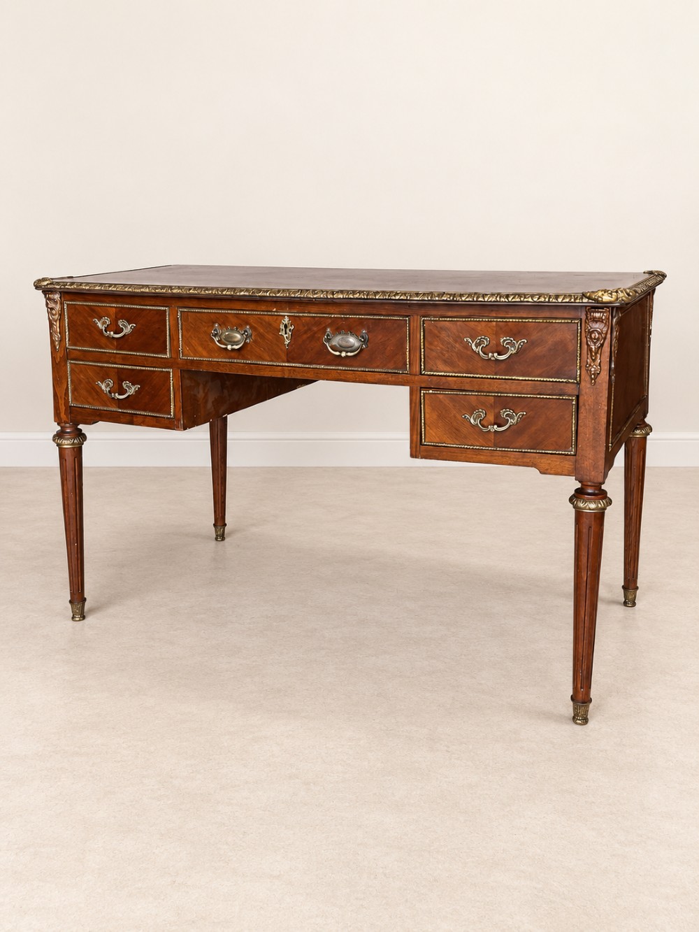 A French Louis XVI style mahogany writing desk featuring a brown leather top, five drawers with cast brass handles, and gilt bronze mounts on fluted tapering legs.