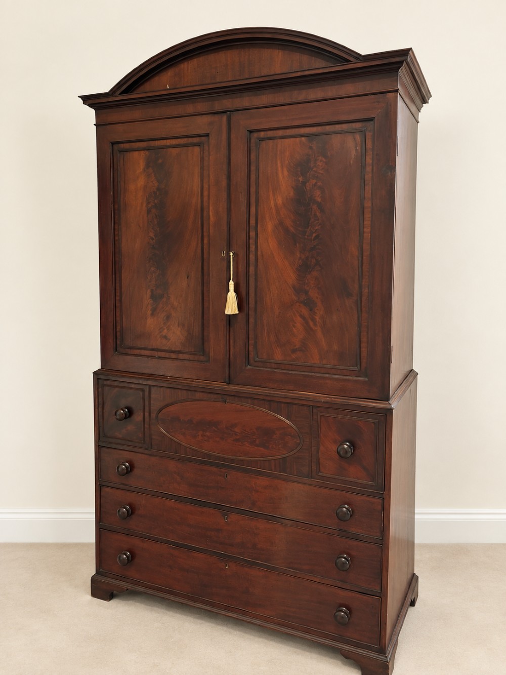An antique Georgian mahogany linen press featuring an arched pediment, twin cupboard doors with flame mahogany panels, and a base chest with multiple drawers.