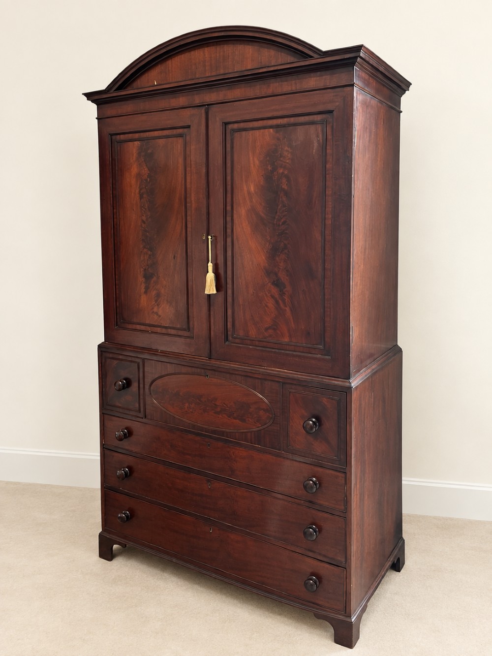 An antique Georgian mahogany linen press featuring an arched pediment, twin cupboard doors with flame mahogany panels, and a base chest with multiple drawers. View II