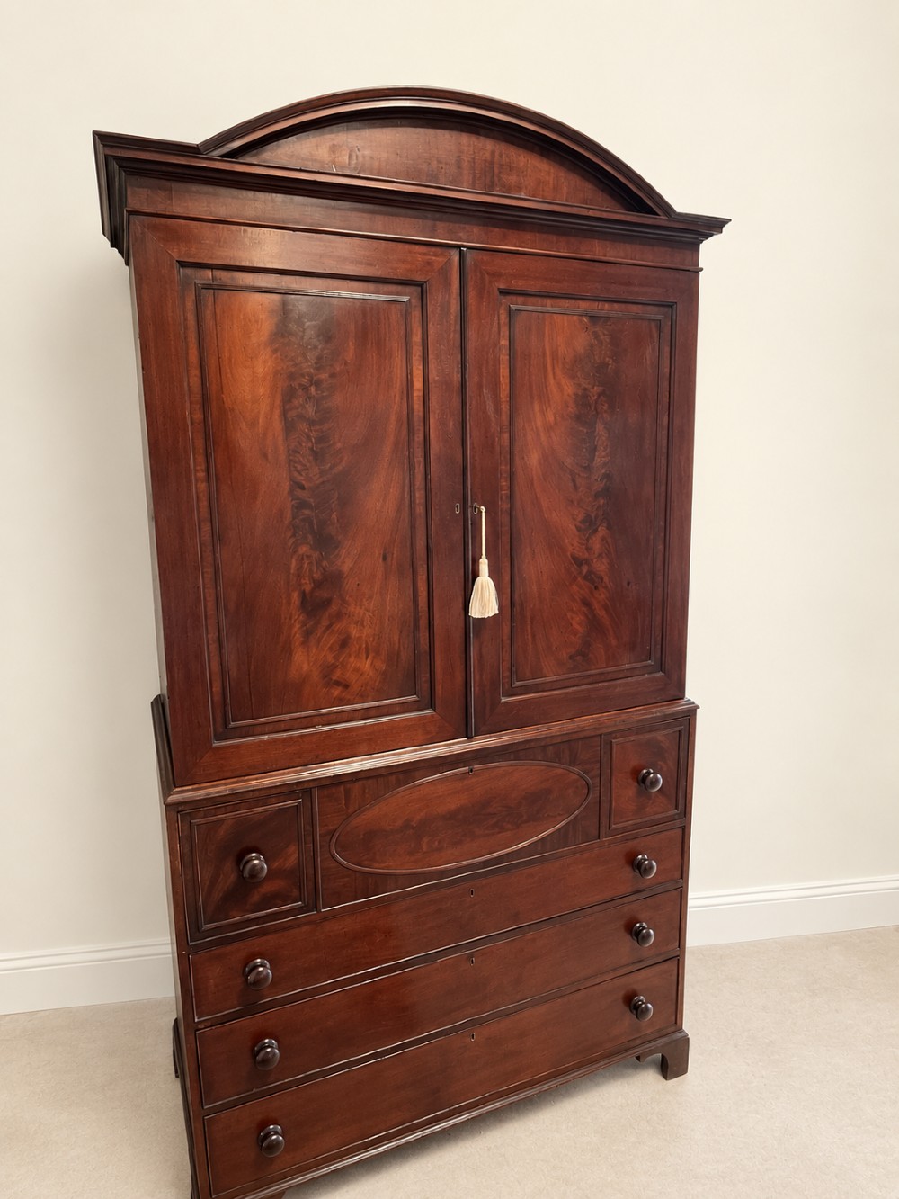 An antique Georgian mahogany linen press featuring an arched pediment, twin cupboard doors with flame mahogany panels, and a base chest with multiple drawers. View III