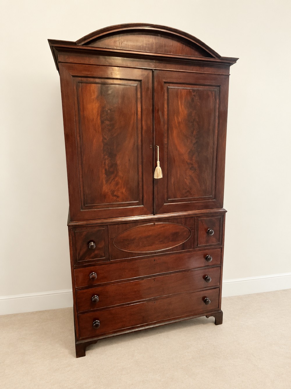 An antique Georgian mahogany linen press featuring an arched pediment, twin cupboard doors with flame mahogany panels, and a base chest with multiple drawers. View IV