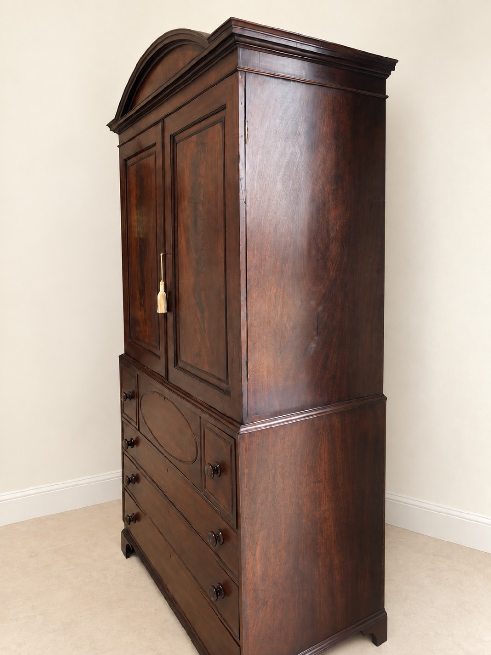 An antique Georgian mahogany linen press featuring an arched pediment, twin cupboard doors with flame mahogany panels, and a base chest with multiple drawers. View VI