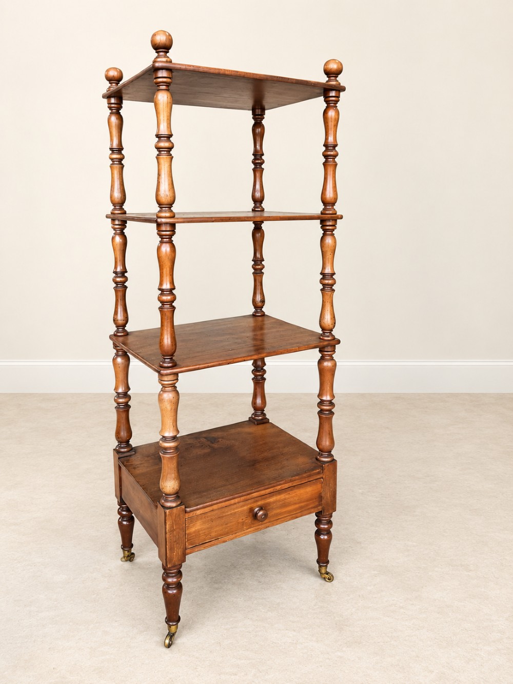 A nineteenth-century mahogany four-tier what-not stand with turned column supports, top finials, a bottom drawer, and brass castors.