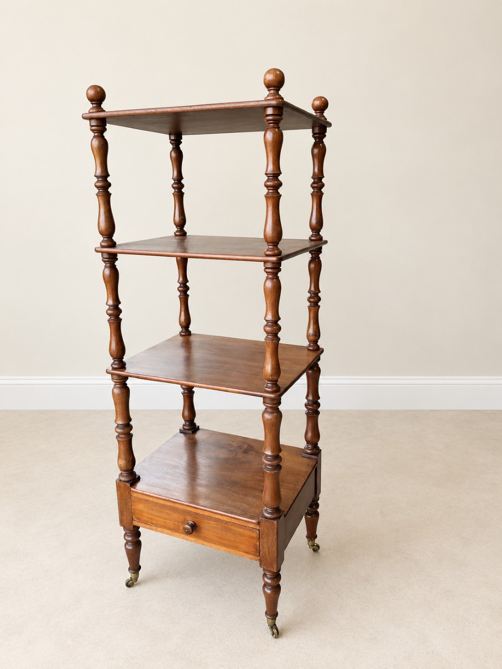 A nineteenth-century mahogany four-tier what-not stand with turned column supports, top finials, a bottom drawer, and brass castors. View II