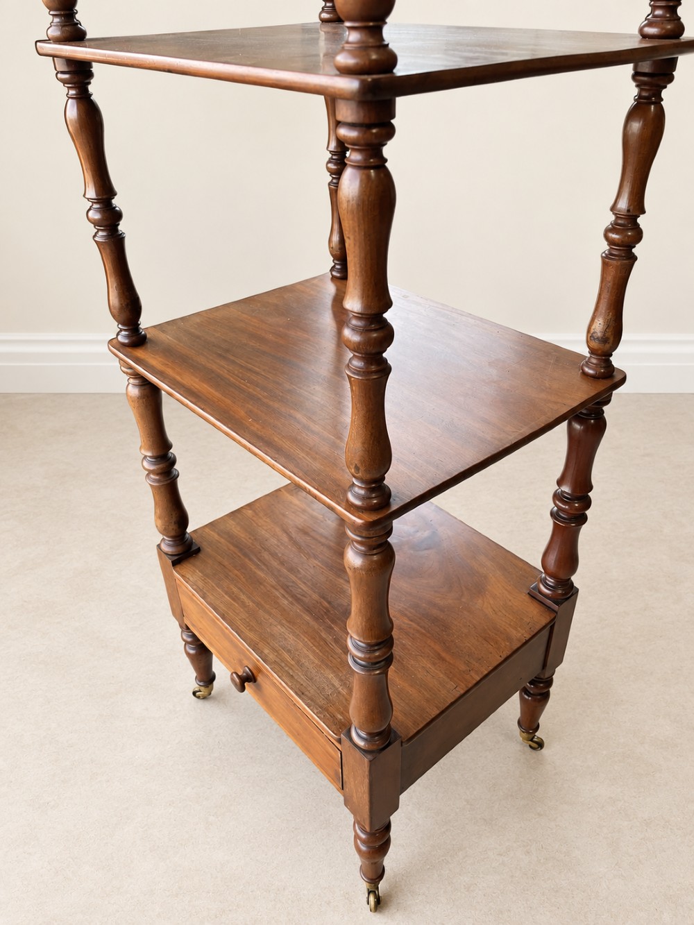 A nineteenth-century mahogany four-tier what-not stand with turned column supports, top finials, a bottom drawer, and brass castors. View IV