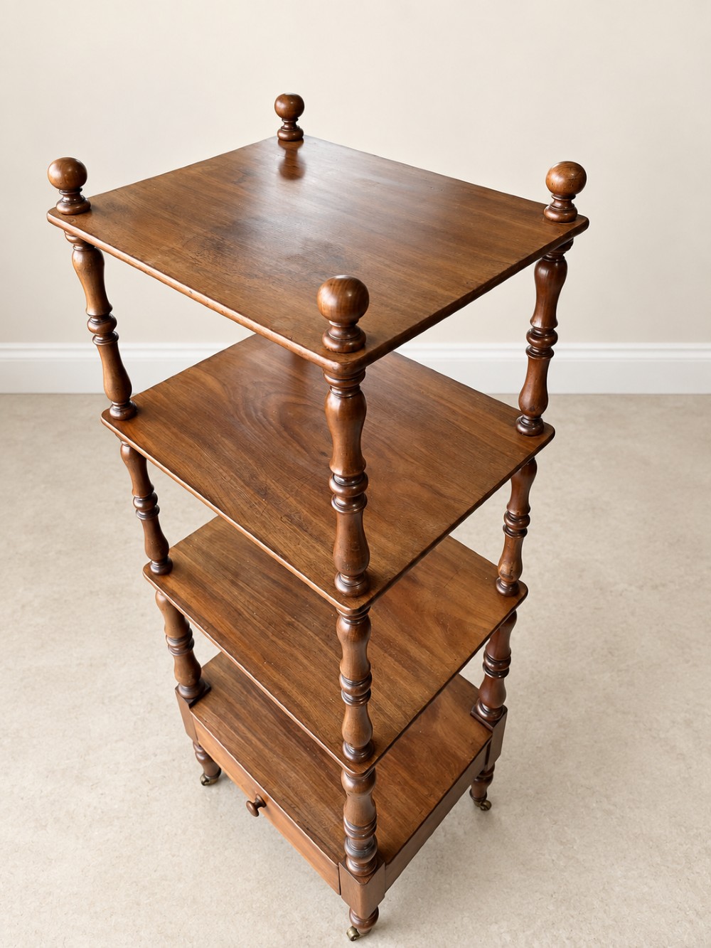 A nineteenth-century mahogany four-tier what-not stand with turned column supports, top finials, a bottom drawer, and brass castors. View V