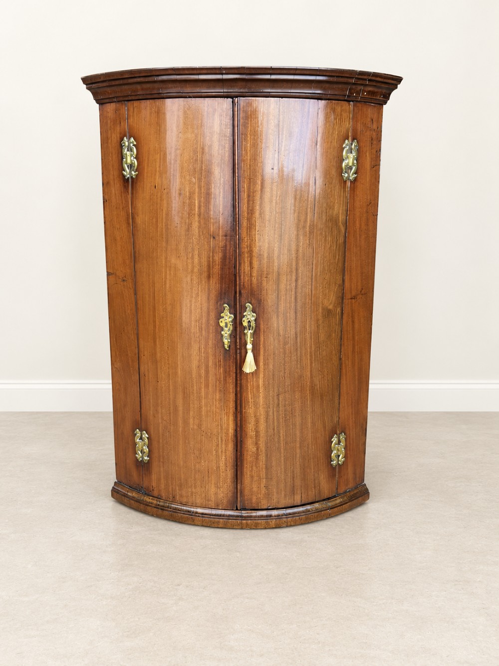An antique Georgian mahogany corner cupboard featuring a curved bowfront double-door structure with ornate brass hinges and moulding.