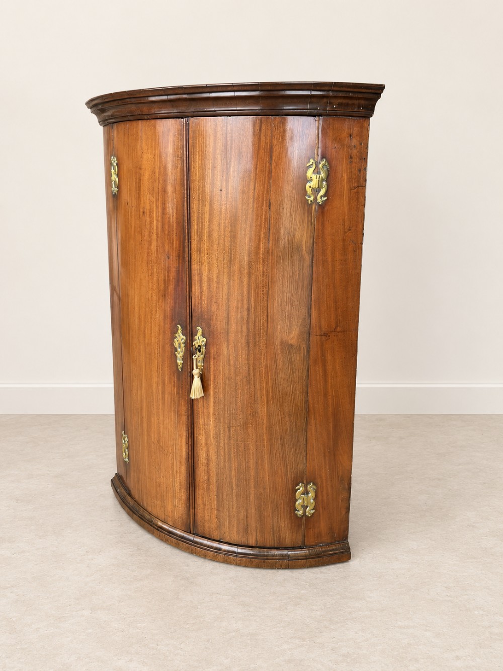 An antique Georgian mahogany corner cupboard featuring a curved bowfront double-door structure with ornate brass hinges and moulding. View II