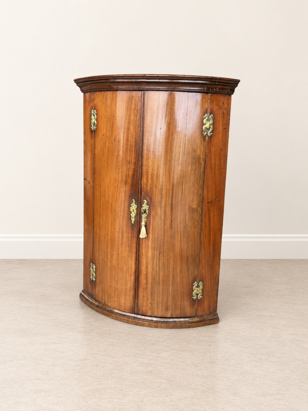An antique Georgian mahogany corner cupboard featuring a curved bowfront double-door structure with ornate brass hinges and moulding. View III