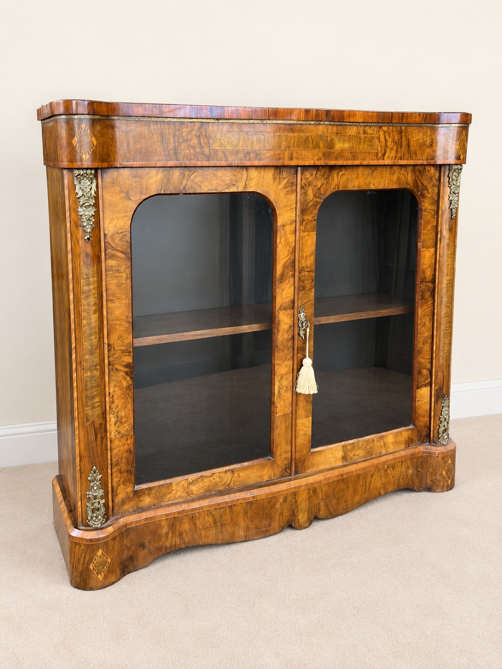 A 19th-century burr walnut low bookcase featuring two glazed doors, a serpentine front profile, geometric marquetry inlay, and decorative brass mounts.