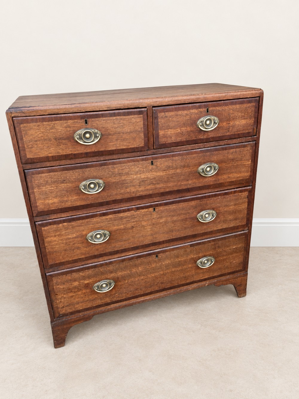 An antique Georgian oak chest of drawers featuring two short drawers above three long drawers with oval brass handles and bracket feet. View II