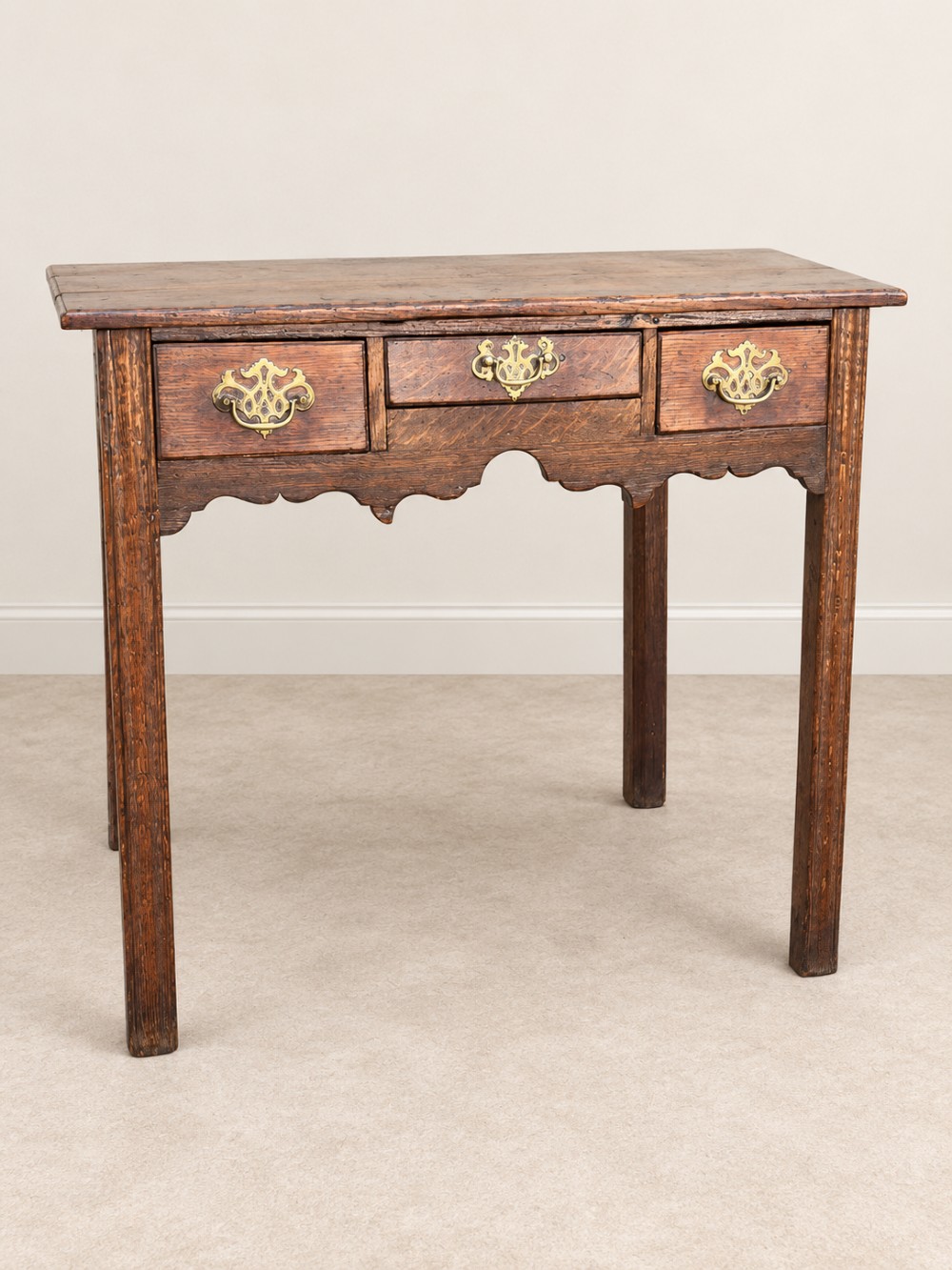 An 18th-century dark oak lowboy side table featuring three drawers with brass handles and a scalloped apron.