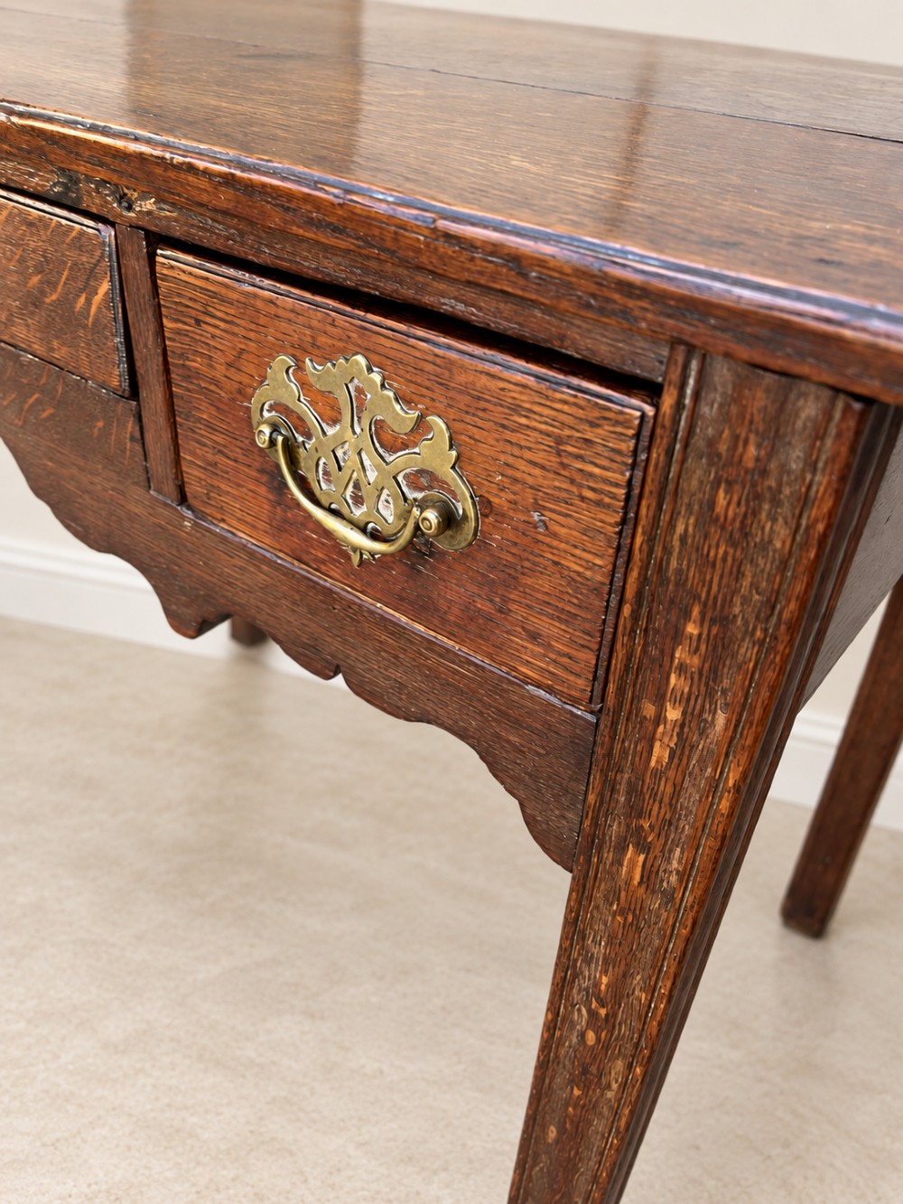 An 18th-century dark oak lowboy side table featuring three drawers with brass handles and a scalloped apron. View V