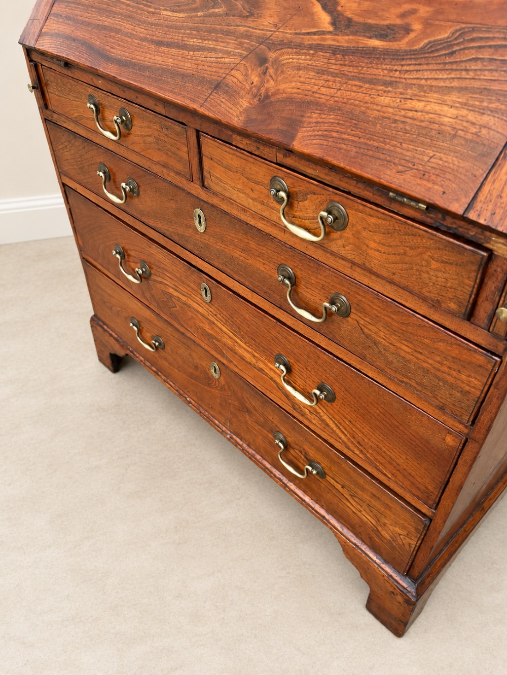 An antique Georgian warm brown elm slant-front writing bureau with four drawers, brass handles, and bracket feet. View X