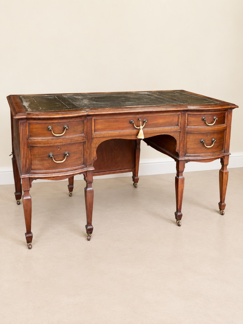 Late nineteenth-century mahogany writing desk featuring an inset dark leather top, five drawers with brass swan-neck handles, fluted pilasters, and tapered legs with brass castors. View IV