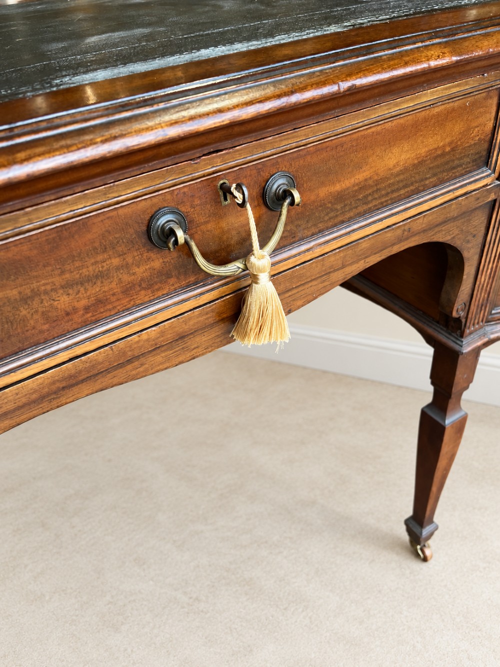 Late nineteenth-century mahogany writing desk featuring an inset dark leather top, five drawers with brass swan-neck handles, fluted pilasters, and tapered legs with brass castors. View VI