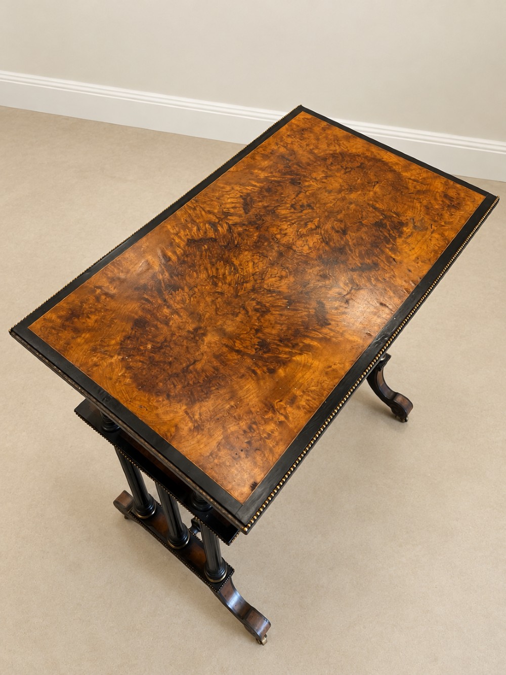A 19th-century two-tier side table with a burr walnut surface, ebonised turned column legs, brass beaded borders, and scrolled feet on castors. View V