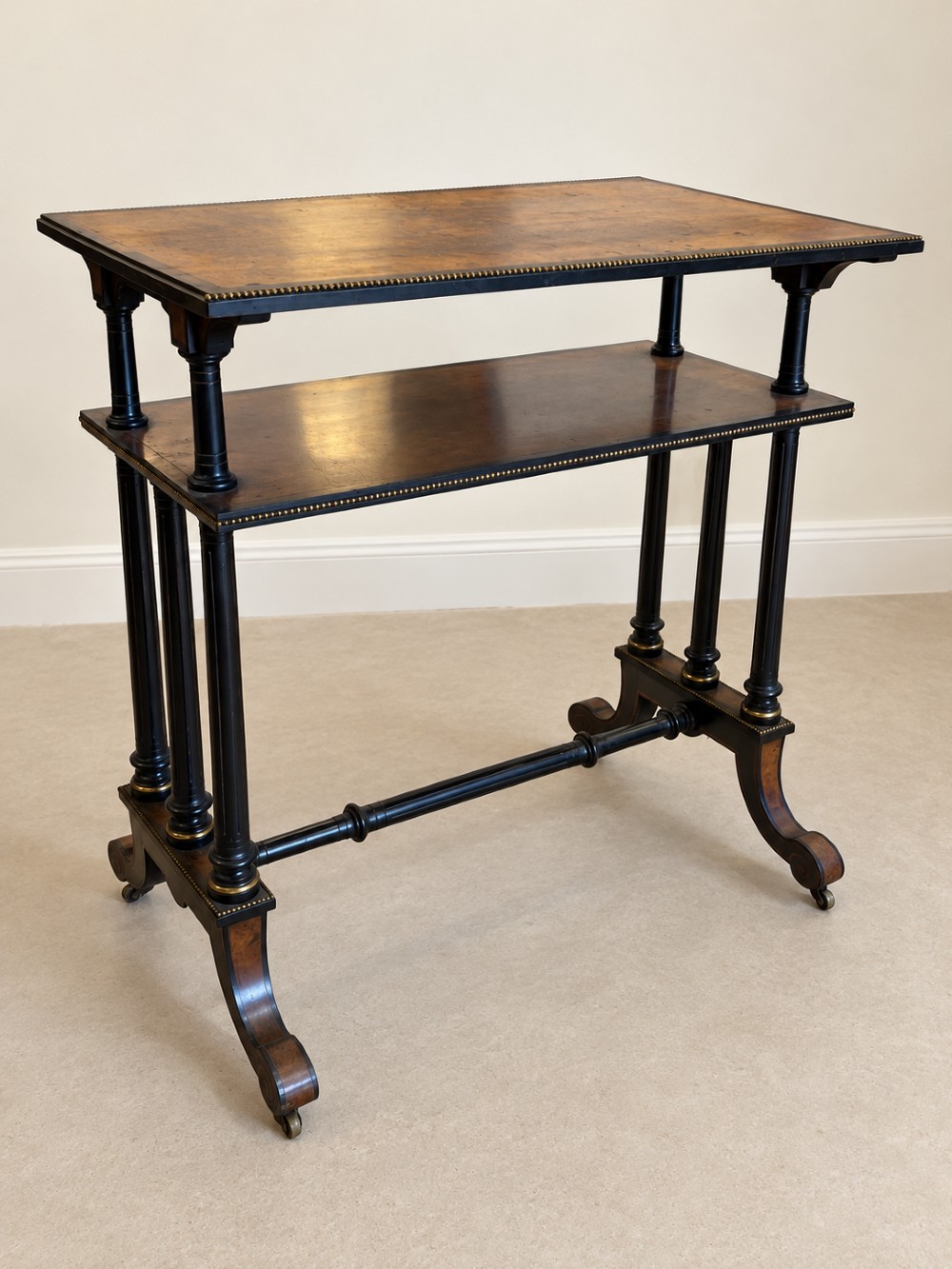 A 19th-century two-tier side table with a burr walnut surface, ebonised turned column legs, brass beaded borders, and scrolled feet on castors. View VI