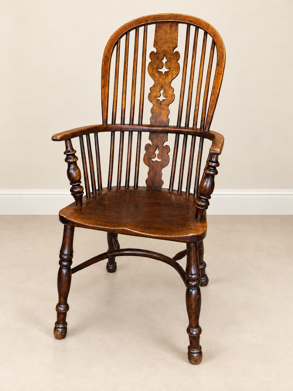 An antique 19th-century wooden Windsor armchair featuring a hoop back with turned spindles, a central pierced splat, a contoured saddle seat, and curved armrests supported by turned front legs.
