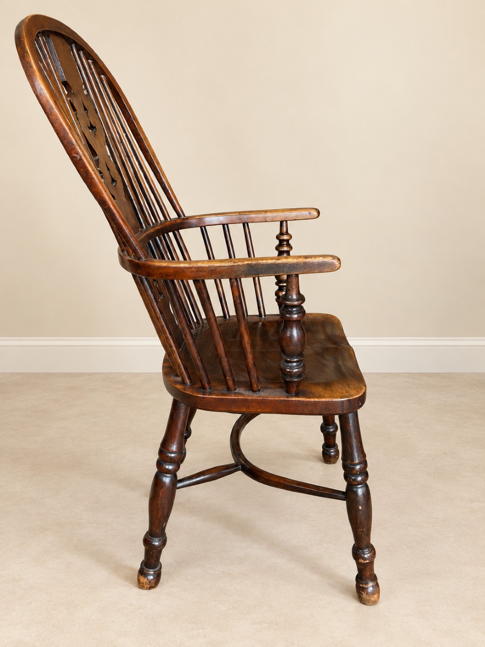 An antique 19th-century wooden Windsor armchair featuring a hoop back with turned spindles, a central pierced splat, a contoured saddle seat, and curved armrests supported by turned front legs. View X
