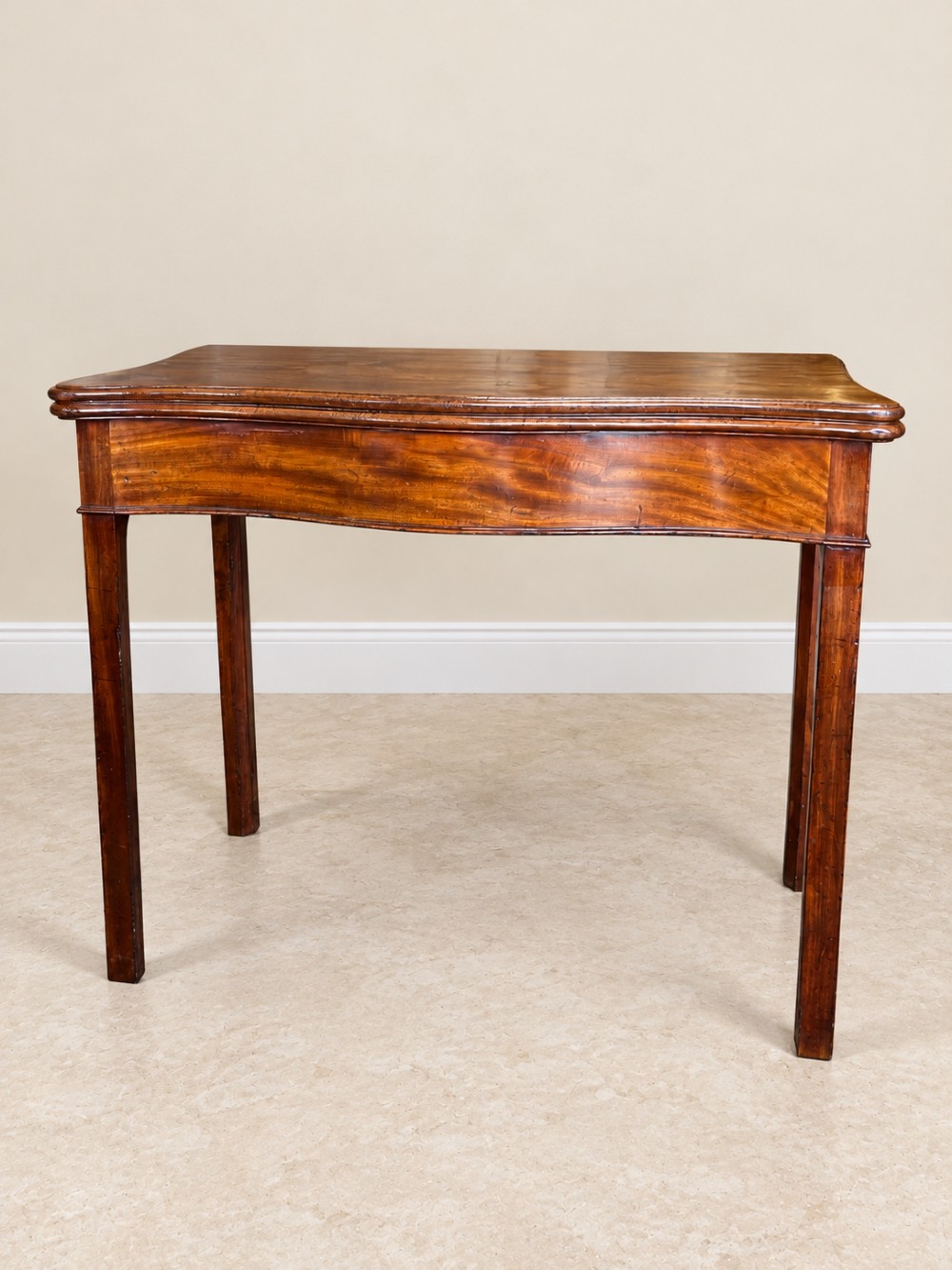 An antique Georgian dark mahogany fold-over table featuring a serpentine-curved front apron and four straight legs.