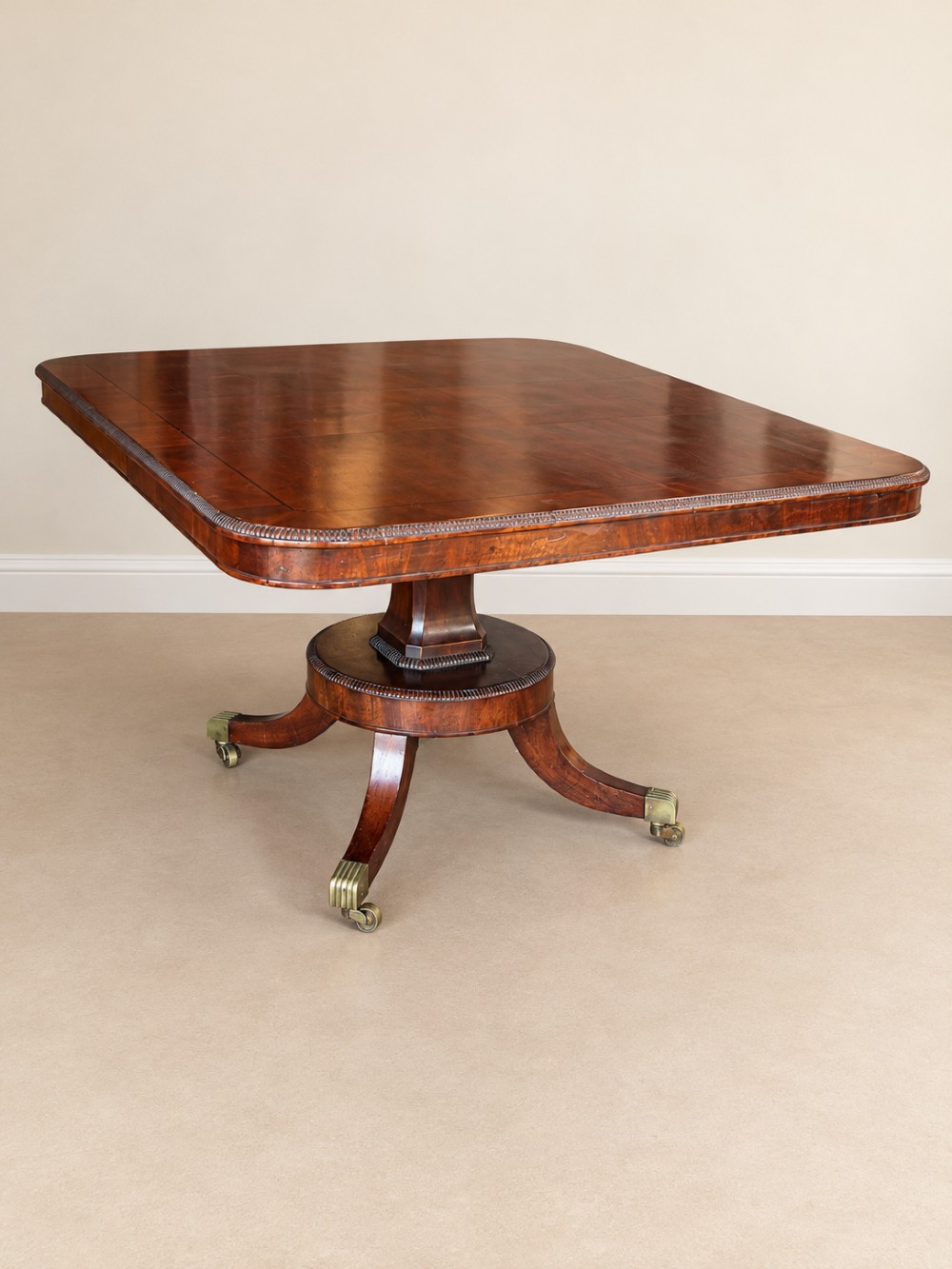 A Regency period mahogany dining table featuring a square top with rounded corners, supported by a central pedestal and four splayed legs with brass castors.