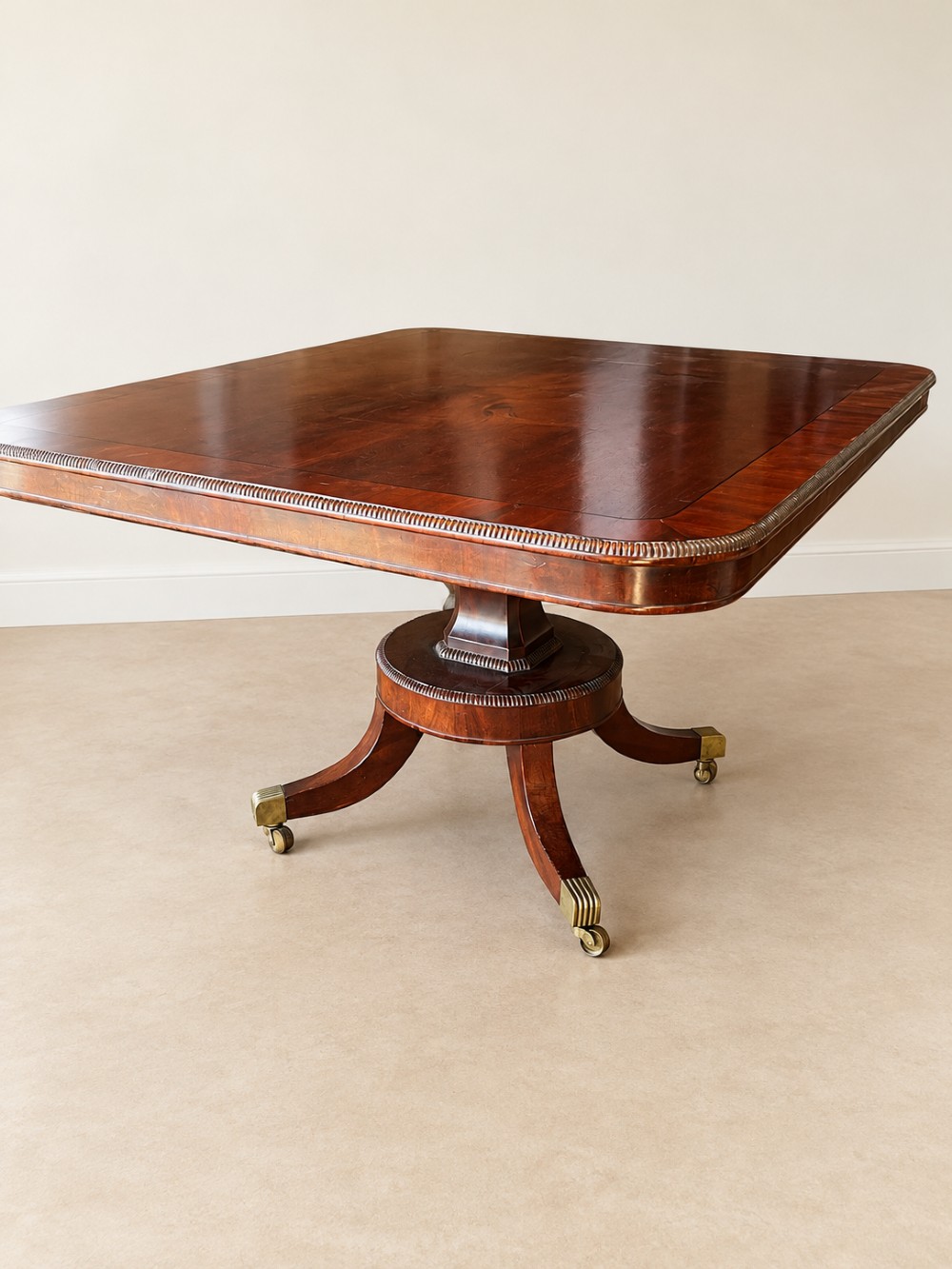 A Regency period mahogany dining table featuring a square top with rounded corners, supported by a central pedestal and four splayed legs with brass castors. View II
