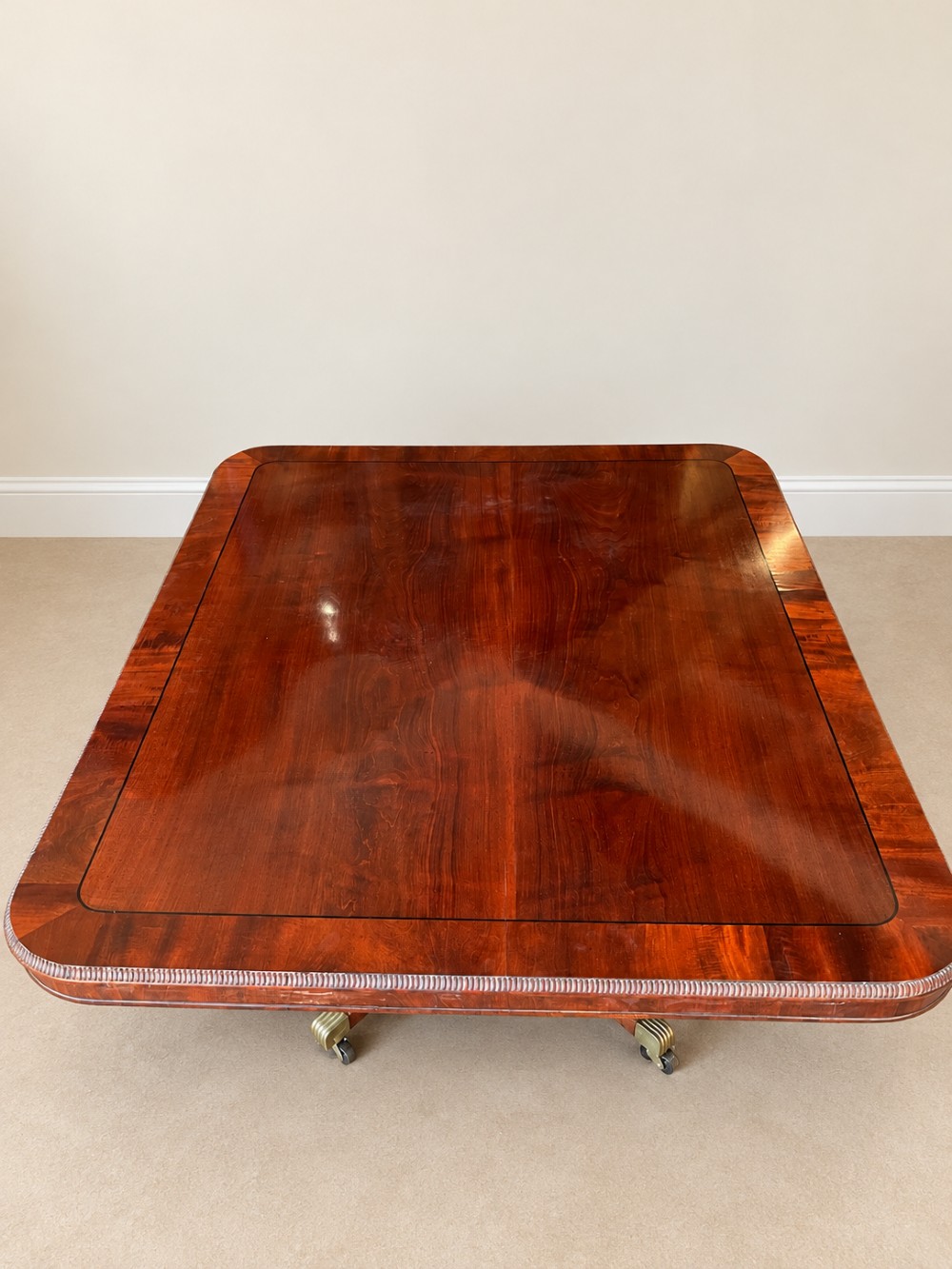 A Regency period mahogany dining table featuring a square top with rounded corners, supported by a central pedestal and four splayed legs with brass castors. View IV