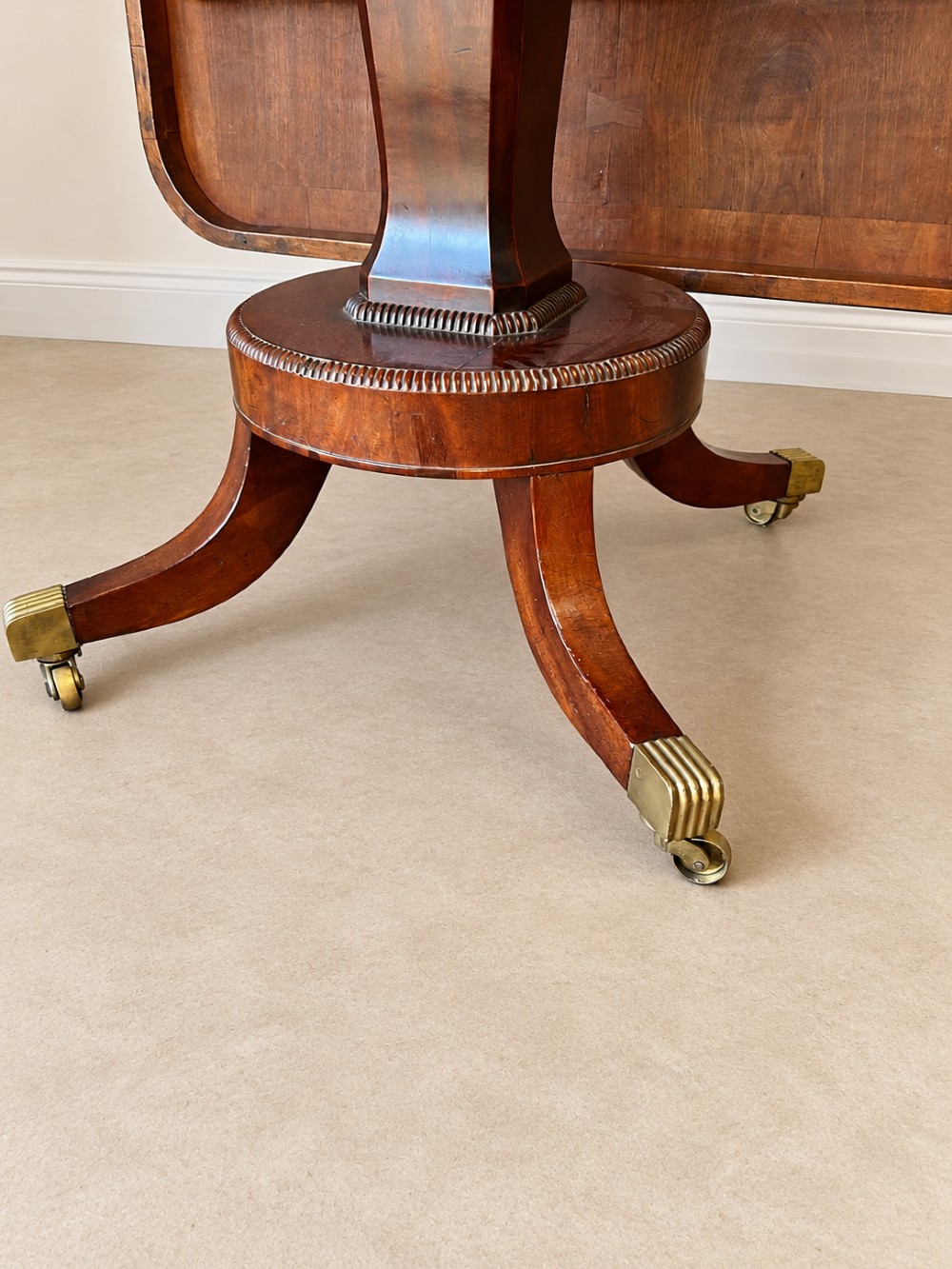 A Regency period mahogany dining table featuring a square top with rounded corners, supported by a central pedestal and four splayed legs with brass castors. View VI