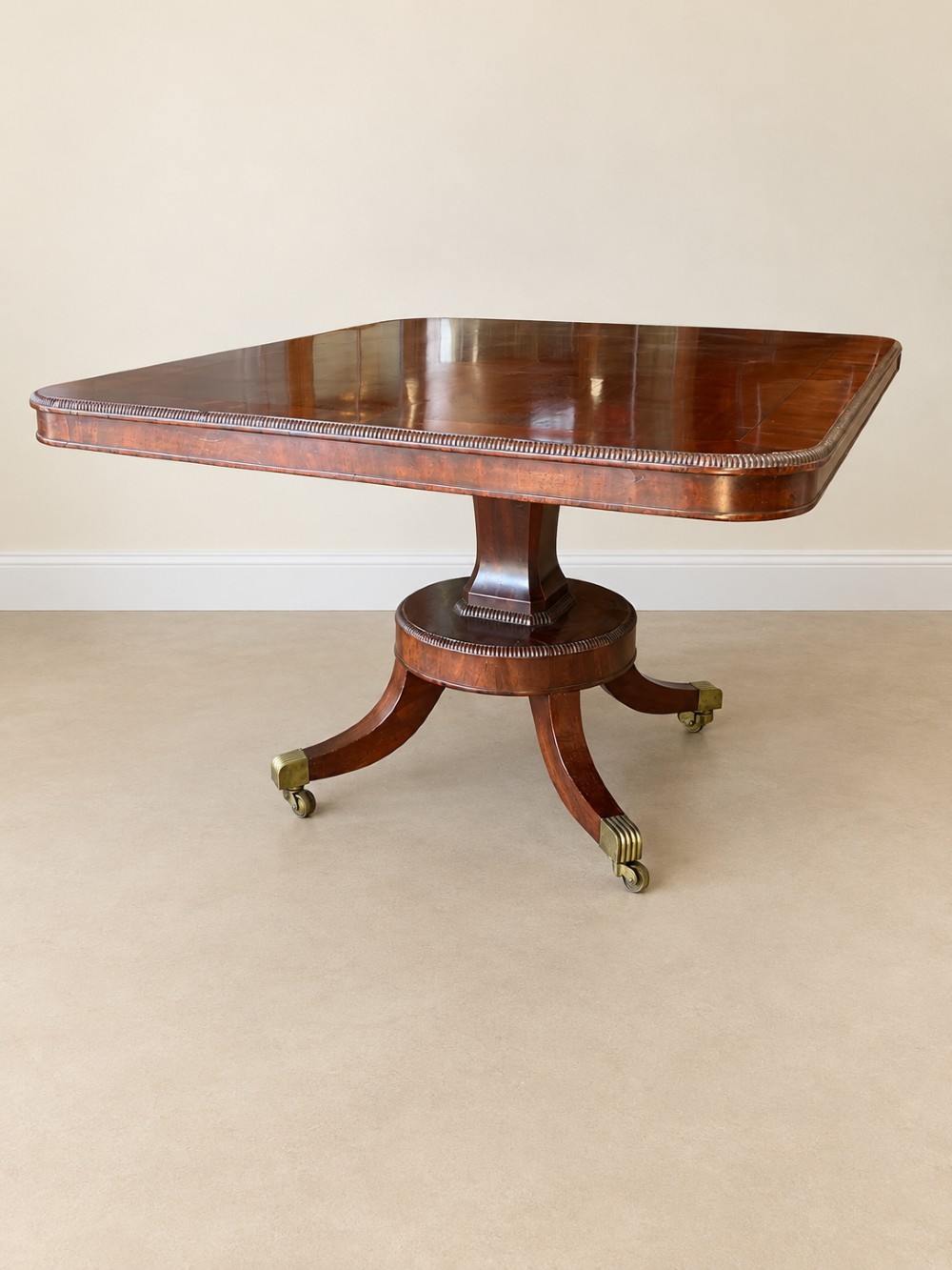 A Regency period mahogany dining table featuring a square top with rounded corners, supported by a central pedestal and four splayed legs with brass castors. View VII