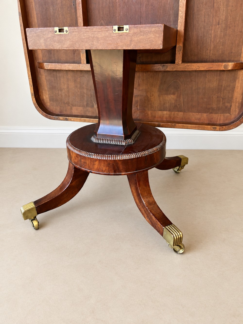 A Regency period mahogany dining table featuring a square top with rounded corners, supported by a central pedestal and four splayed legs with brass castors. View VIII